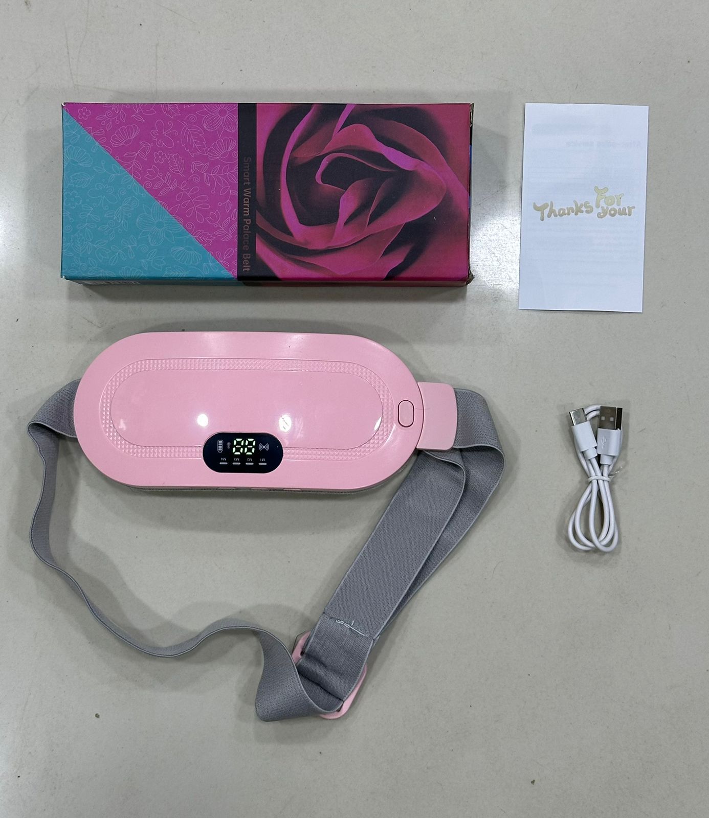 Pink electronic device with gray strap, box, and USB cable on a light gray surface